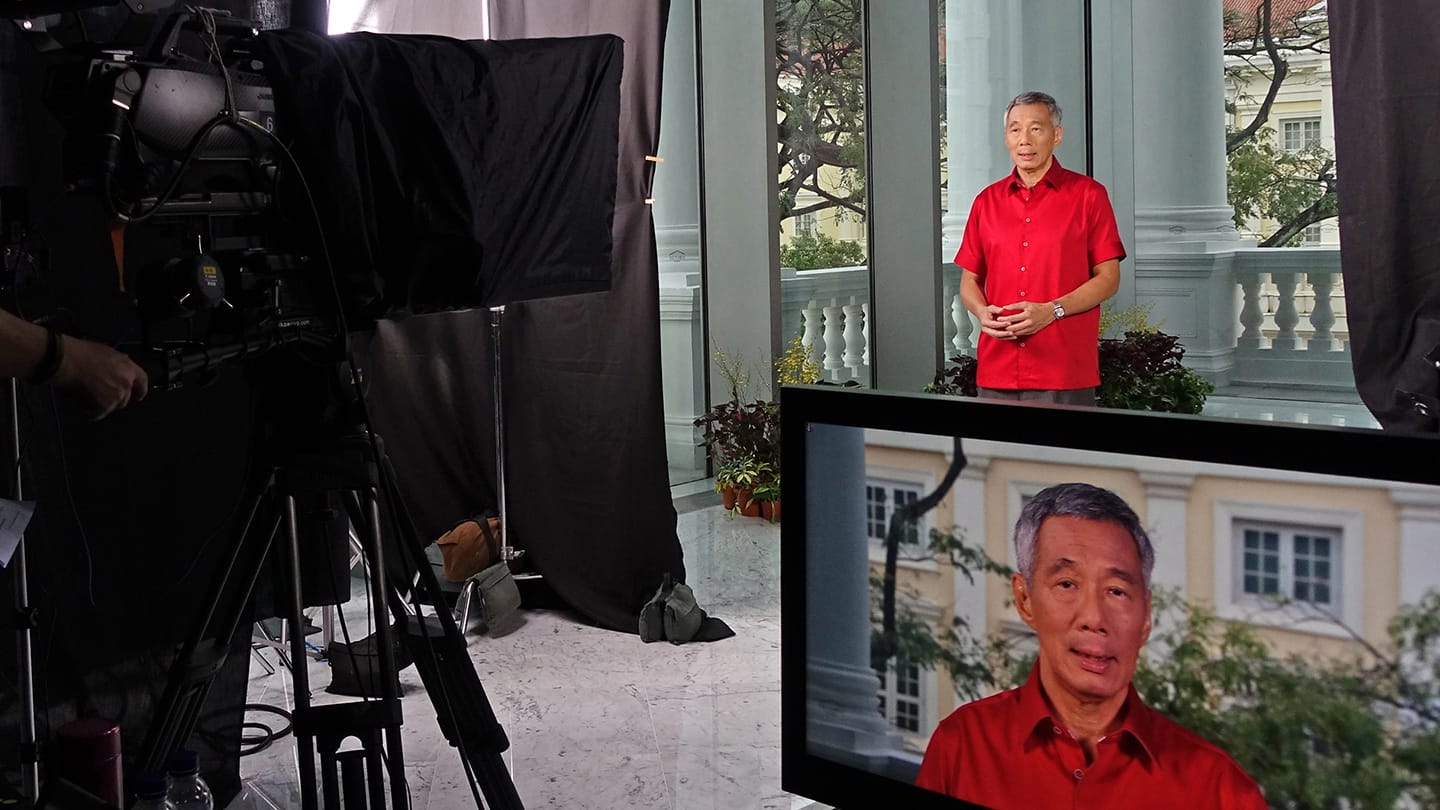Lee Hsien Loong on set in a red shirt, with filming equipment and a monitor showing his face.